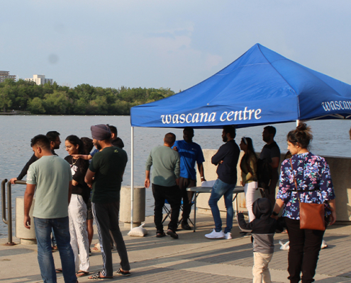 People stand around a blue tent with the words "Wascana Centre: on it. They are by a lake.