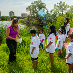 Photo of a group of kids wearing white t-shirts, one is holding a blue net. A womanstands in front of them wearing a pink shirt holding a container. The scenary around them is nature.