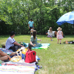 People sitting around on a blanket on the grass, surrounded by trees in the distance. There is a blue umbrella set up.