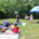 People sitting around on a blanket on the grass, surrounded by trees in the distance. There is a blue umbrella set up.