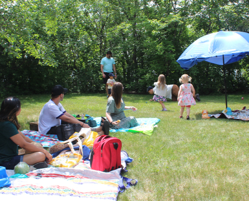 People sitting around on a blanket on the grass, surrounded by trees in the distance. There is a blue umbrella set up.
