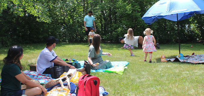 People sitting around on a blanket on the grass, surrounded by trees in the distance. There is a blue umbrella set up.