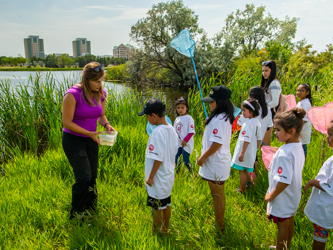 Photo of a group of kids wearing white t-shirts, one is holding a blue net. A womanstands in front of them wearing a pink shirt holding a container. The scenary around them is nature.