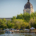 A wide shot of Wascana Lake features a motorized boat on Wascana Lake with people, a white tent with people under it, while the Saskatchewan Legislative Building peeks behind a wall of green trees.