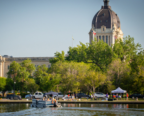 A wide shot of Wascana Lake features a motorized boat on Wascana Lake with people, a white tent with people under it, while the Saskatchewan Legislative Building peeks behind a wall of green trees.