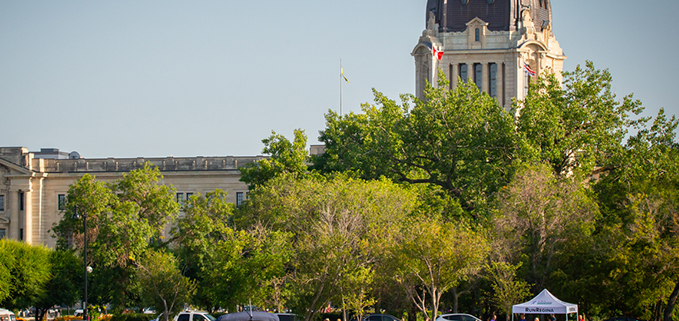 A wide shot of Wascana Lake features a motorized boat on Wascana Lake with people, a white tent with people under it, while the Saskatchewan Legislative Building peeks behind a wall of green trees.