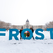 A family stands behind giant blue letters that spell out "FROST". The Saskatchewan Legislative Building is in the background.