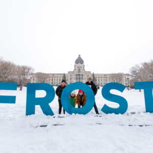 A family stands behind giant blue letters that spell out "FROST". The Saskatchewan Legislative Building is in the background.