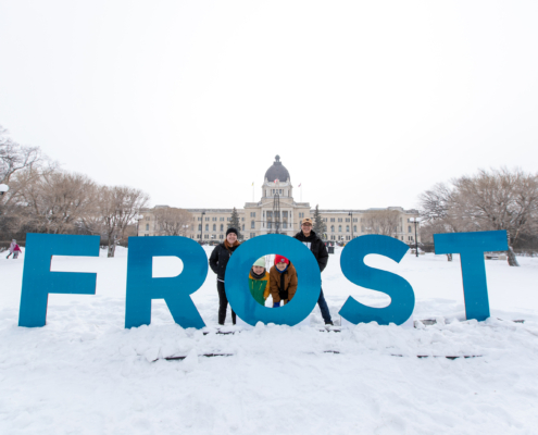 A family stands behind giant blue letters that spell out "FROST". The Saskatchewan Legislative Building is in the background.