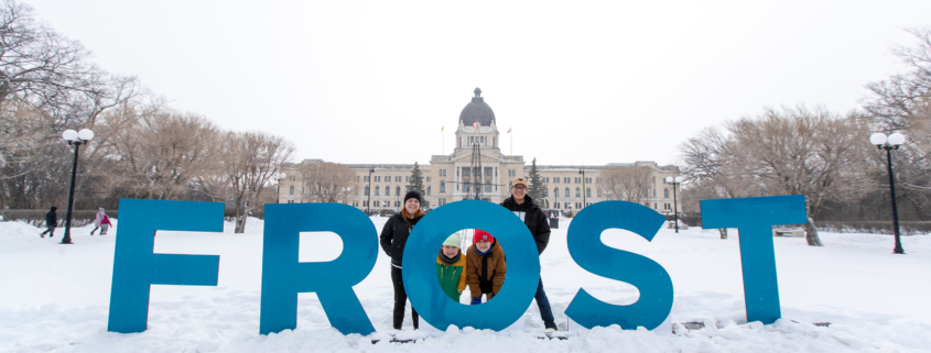 A family stands behind giant blue letters that spell out "FROST". The Saskatchewan Legislative Building is in the background.
