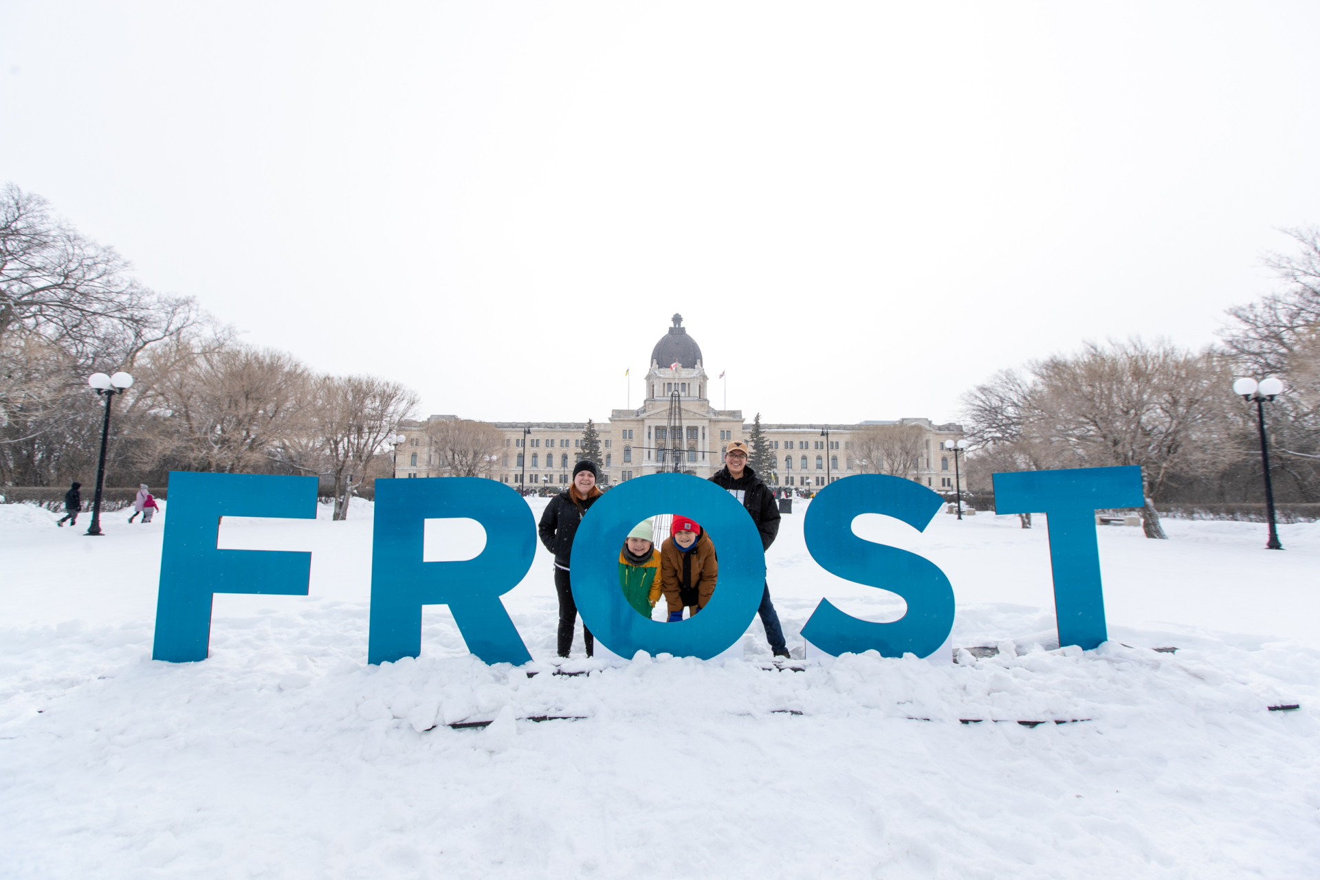 A family stands behind giant blue letters that spell out "FROST". The Saskatchewan Legislative Building is in the background.