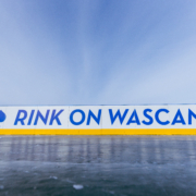A close-up view of an outdoor ice rink barrier with the words “RINK ON WASCANA” in bold blue letters on a white background, accented by a yellow strip along the bottom. A blue leaf icon appears to the left of the text. The sky above is clear and bright, creating a crisp winter atmosphere.