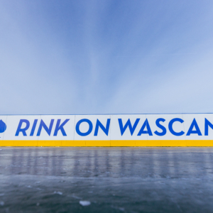 A close-up view of an outdoor ice rink barrier with the words “RINK ON WASCANA” in bold blue letters on a white background, accented by a yellow strip along the bottom. A blue leaf icon appears to the left of the text. The sky above is clear and bright, creating a crisp winter atmosphere.