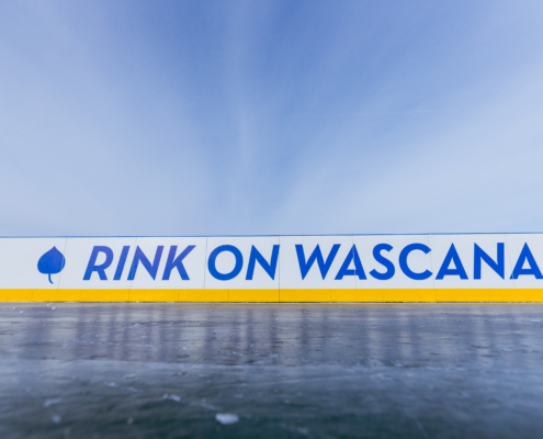 A close-up view of an outdoor ice rink barrier with the words “RINK ON WASCANA” in bold blue letters on a white background, accented by a yellow strip along the bottom. A blue leaf icon appears to the left of the text. The sky above is clear and bright, creating a crisp winter atmosphere.