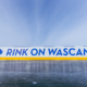 A close-up view of an outdoor ice rink barrier with the words “RINK ON WASCANA” in bold blue letters on a white background, accented by a yellow strip along the bottom. A blue leaf icon appears to the left of the text. The sky above is clear and bright, creating a crisp winter atmosphere.