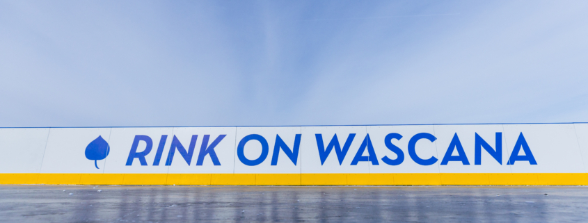 A close-up view of an outdoor ice rink barrier with the words “RINK ON WASCANA” in bold blue letters on a white background, accented by a yellow strip along the bottom. A blue leaf icon appears to the left of the text. The sky above is clear and bright, creating a crisp winter atmosphere.