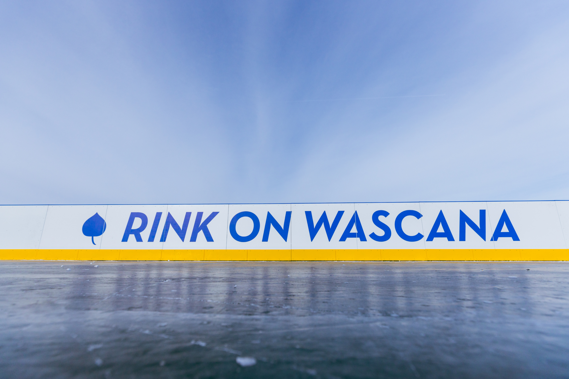 A close-up view of an outdoor ice rink barrier with the words “RINK ON WASCANA” in bold blue letters on a white background, accented by a yellow strip along the bottom. A blue leaf icon appears to the left of the text. The sky above is clear and bright, creating a crisp winter atmosphere.