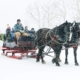 Two black horses pulling a red sleigh through falling snow with passengers seated inside.